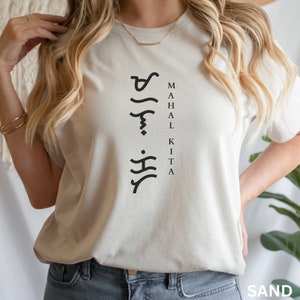 May include: A sand-coloured short-sleeve t-shirt featuring black text that reads "MAHAL KITA" vertically alongside a column of black Baybayin script characters. The shirt has a crew neckline and a standard fit, presented in a neutral tone.