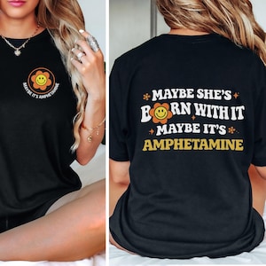 May include: Black t-shirt with a retro design. The front features a smiling flower graphic with the text "MAYBE IT'S AMPHETAMINE". The back of the shirt displays the phrase "MAYBE SHE'S BORN WITH IT MAYBE IT'S AMPHETAMINE" in white and yellow.