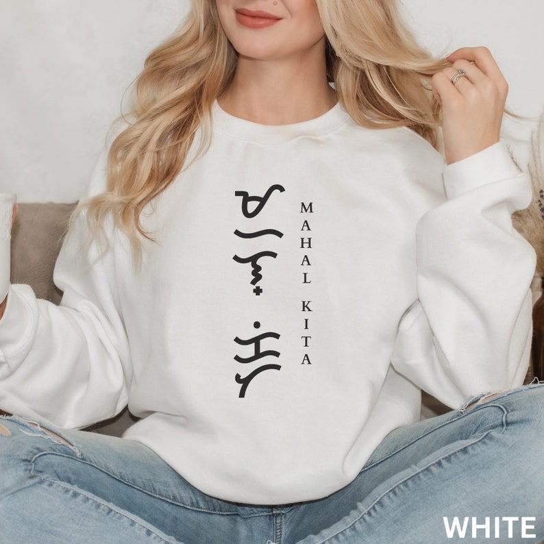 May include: A white long-sleeve crewneck sweatshirt featuring black text that reads "MAHAL KITA" vertically alongside a column of black Baybayin script characters. The garment has a standard fit with ribbed cuffs and a ribbed neckline.