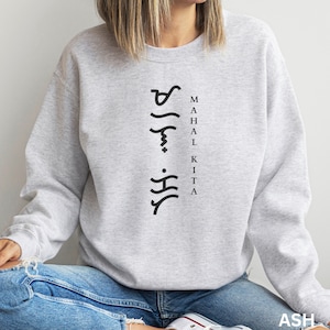 May include: A light grey crewneck sweatshirt featuring black text that reads MAHAL KITA vertically, accompanied by a column of black Baybayin script characters to the left of the lettering. The garment has long sleeves and a ribbed neckline.