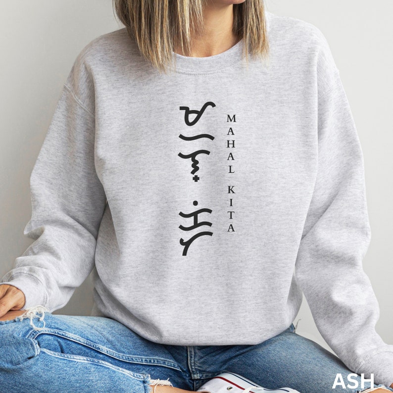May include: A light grey crewneck sweatshirt featuring black text that reads MAHAL KITA vertically, accompanied by a column of black Baybayin script characters to the left of the lettering. The garment has long sleeves and a ribbed neckline.