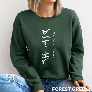 May include: A forest green crewneck sweatshirt featuring white text that reads MAHAL KITA vertically, accompanied by stylised Baybayin script characters arranged in a vertical column to the left of the English lettering.