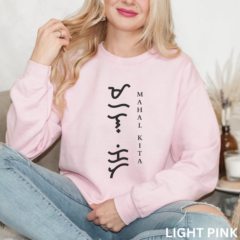 May include: A light pink crewneck sweatshirt featuring black text printed vertically on the chest. The design includes Baybayin script characters arranged in a column next to the words MAHAL KITA in a simple, sans-serif black font.