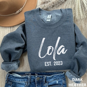 May include: A dark heather grey crewneck sweatshirt featuring the white text Lola and EST. 2023 printed on the front. The garment has long sleeves, a ribbed neckline, and a soft fleece interior lining visible at the cuffs and collar area.