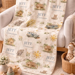 May include: Cream-colored baby blanket with a repeating pattern of cartoon vehicles, huts, suns, and animals. The name "JETT Camansa" is printed in a soft blue font throughout the design. The blanket is draped over a white armchair.