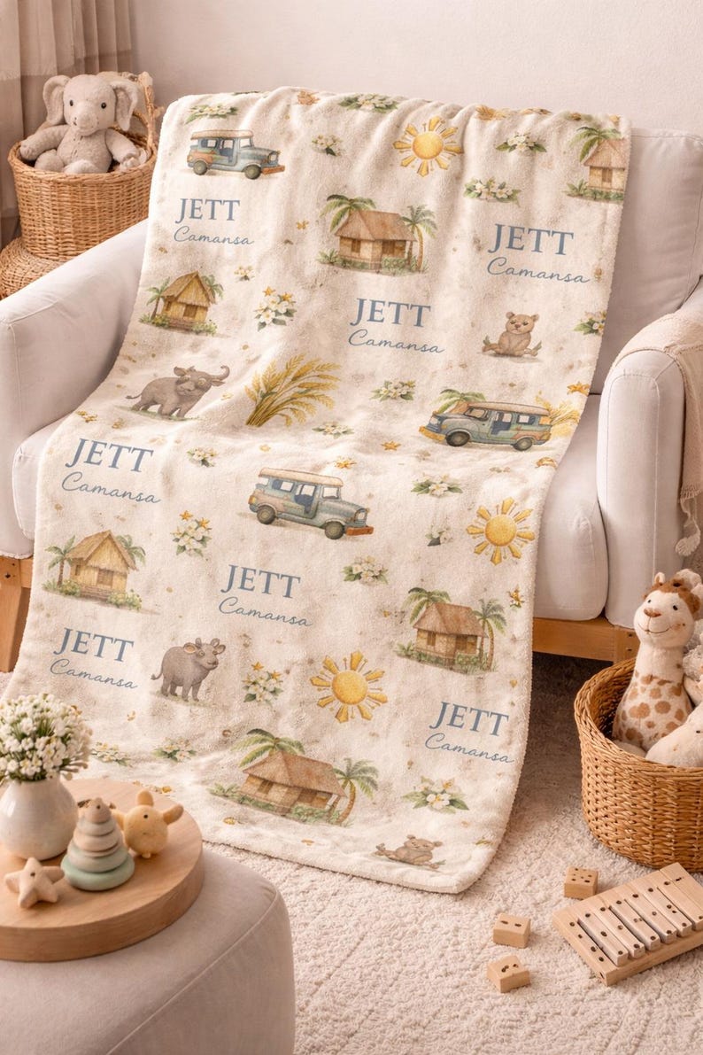 May include: Cream-colored baby blanket with a repeating pattern of cartoon vehicles, huts, suns, and animals. The name "JETT Camansa" is printed in a soft blue font throughout the design. The blanket is draped over a white armchair.