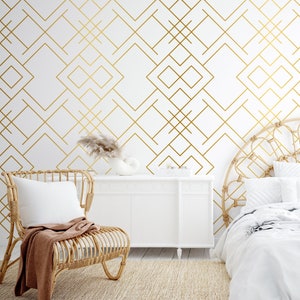 Può includere: Una parete bianca con un motivo geometrico ripetitivo di sottili linee dorate. La stanza include una sedia in rattan di colore chiaro con un cuscino bianco e una coperta marrone, una credenza bianca e un letto con testiera in rattan su un tappeto.