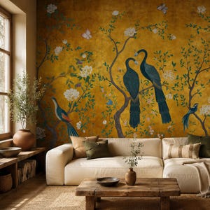 Peut inclure: Une peinture murale sur fond jaune moutarde profond avec des paons bleu sarcelle, de petits oiseaux bleus, des fleurs blanches et des papillons sur des branches brunes. La peinture est située derrière un canapé crème avec des coussins.