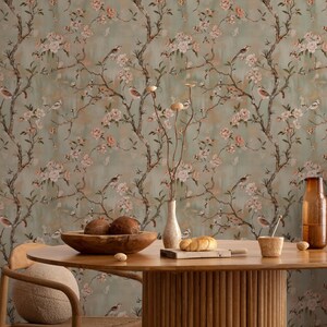 May include: A room with floral wallpaper featuring branches, birds, and pink blossoms. A wooden table with a bowl of fruit, a vase, and other decorative items is in front of the wallpaper. The room has a warm, inviting atmosphere.