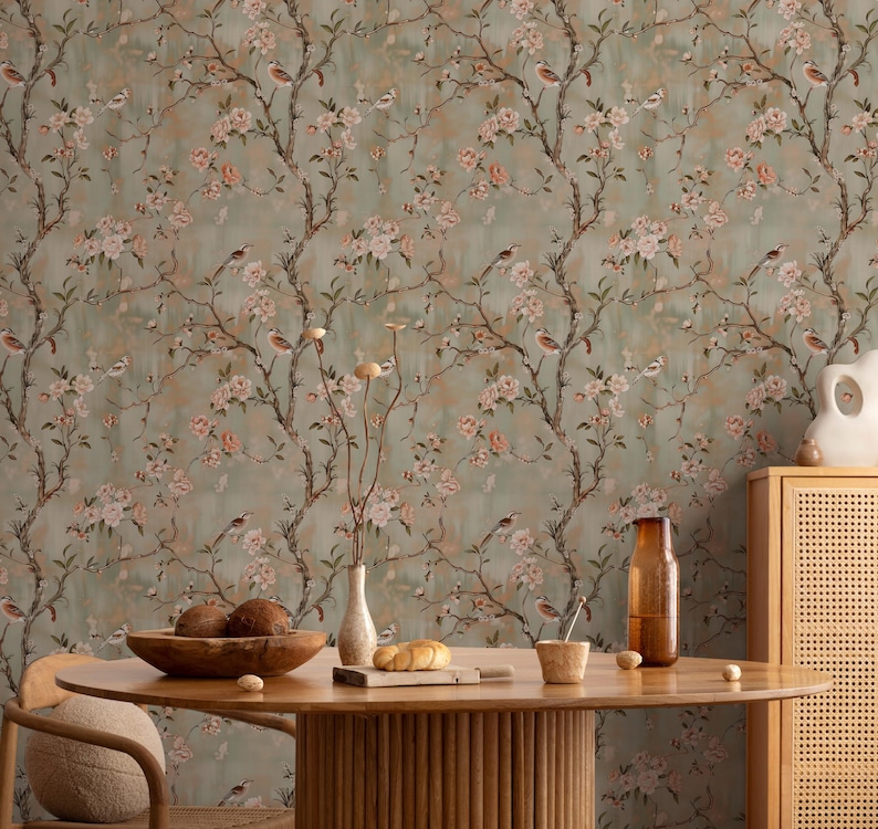 May include: A room with floral wallpaper featuring branches, birds, and pink blossoms. A wooden table with a bowl of fruit, a vase, and other decorative items is in front of the wallpaper. The room has a warm, inviting atmosphere.