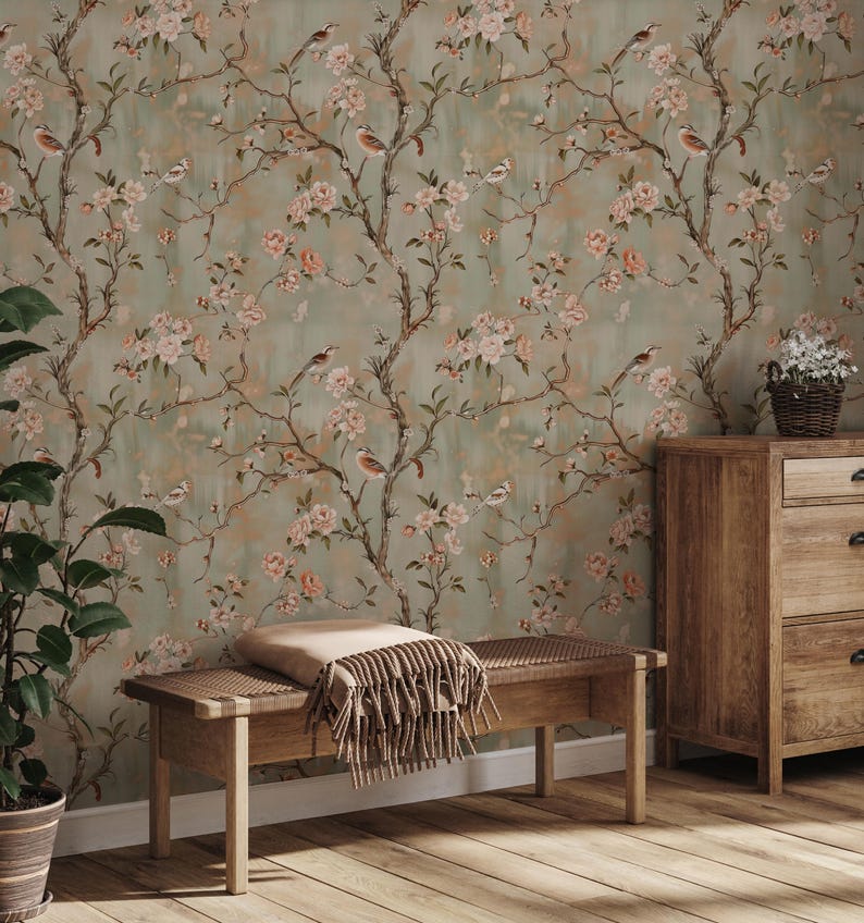 May include: A room with floral wallpaper featuring pink blossoms, birds, and branches on a muted green background. A wooden bench with a blanket and a wooden dresser with a basket of flowers are in the room. The floor is made of wood.