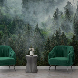 May include: Two green velvet armchairs with black metal legs, a gray round side table with a white vase of flowers, and a gray shelf with a decorative sculpture in front of a green forest wallpaper with fog.