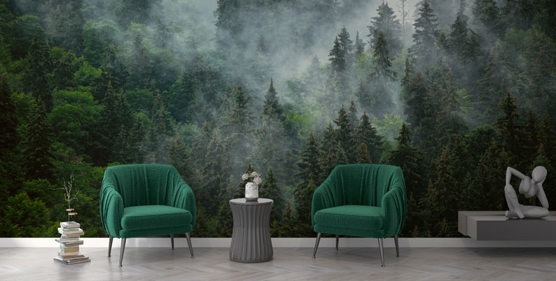May include: Two green velvet armchairs with black metal legs, a gray round side table with a white vase of flowers, and a gray shelf with a decorative sculpture in front of a green forest wallpaper with fog.