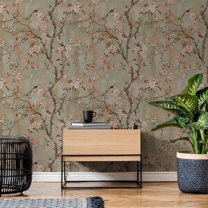 May include: Wallpaper featuring a repeating pattern of brown tree branches with light pink flowers and small birds against a muted sage green background. The design includes delicate floral details and avian figures spread across the surface.