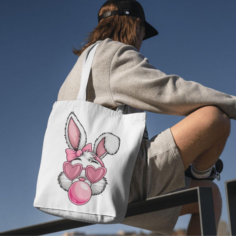 May include: White tote bag with a pink bunny wearing pink heart-shaped sunglasses and blowing a pink bubble. The bunny has a pink bow on its head.