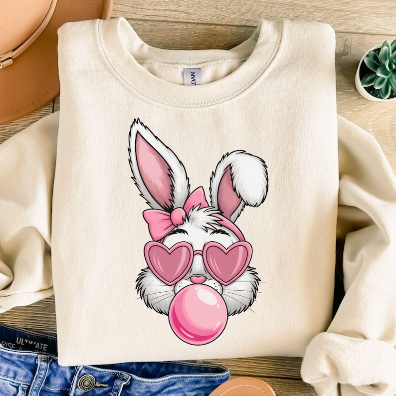 May include: A white bunny with pink ears, a pink bow, and pink heart-shaped sunglasses is blowing a pink bubble. The bunny is on a cream-colored sweatshirt.