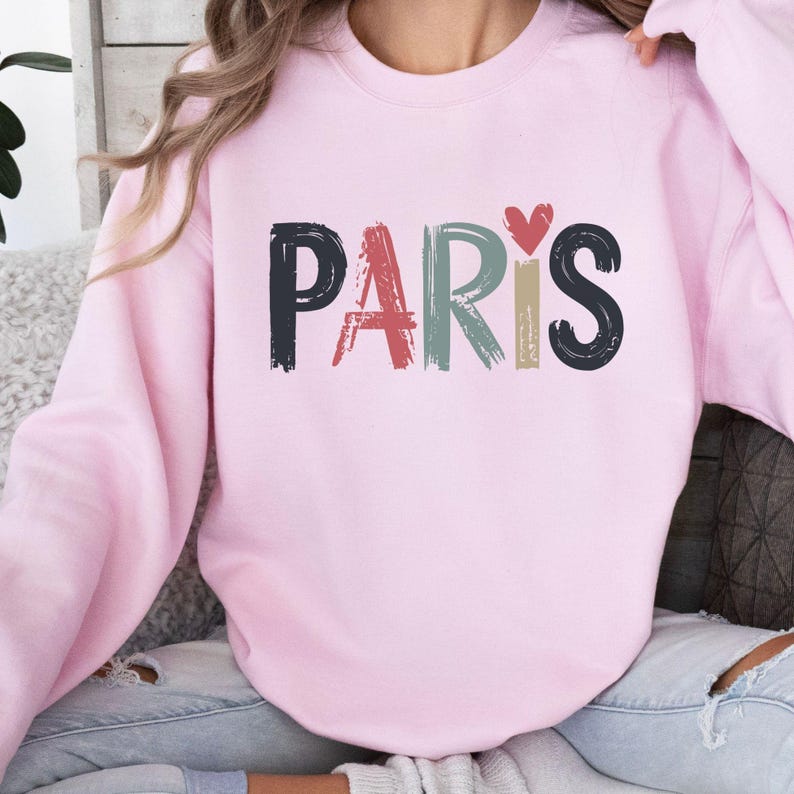 May include: A pink sweatshirt with the word "PARIS" written in colorful, brushstroke letters with a small red heart above the "S".