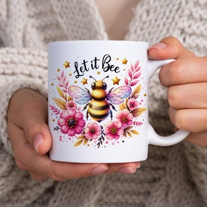 May include: White ceramic mug with a bee and flowers design. The mug has the text "Let it Bee" in black lettering.