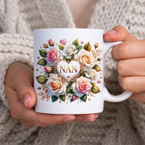 May include: White ceramic mug with a gold heart design featuring the word "NAN" surrounded by pink and white roses and gold hearts.