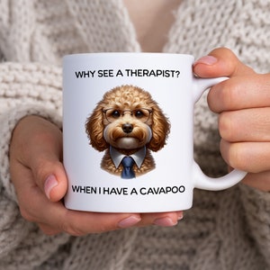 Puede incluir: Taza de cerámica blanca con un perro marrón y blanco que lleva gafas y una corbata azul. La taza dice "¿Por qué ver a un terapeuta? Cuando tengo un cavapoo."