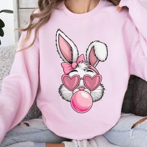 May include: Pink sweatshirt with a white bunny wearing pink heart-shaped sunglasses and blowing a pink bubble. The bunny has a pink bow on its head.