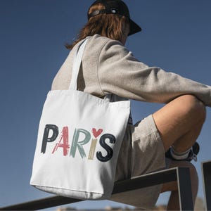 May include: A white tote bag with the word "PARIS" printed in colorful, brushstroke-style lettering. A small red heart is included in the design.