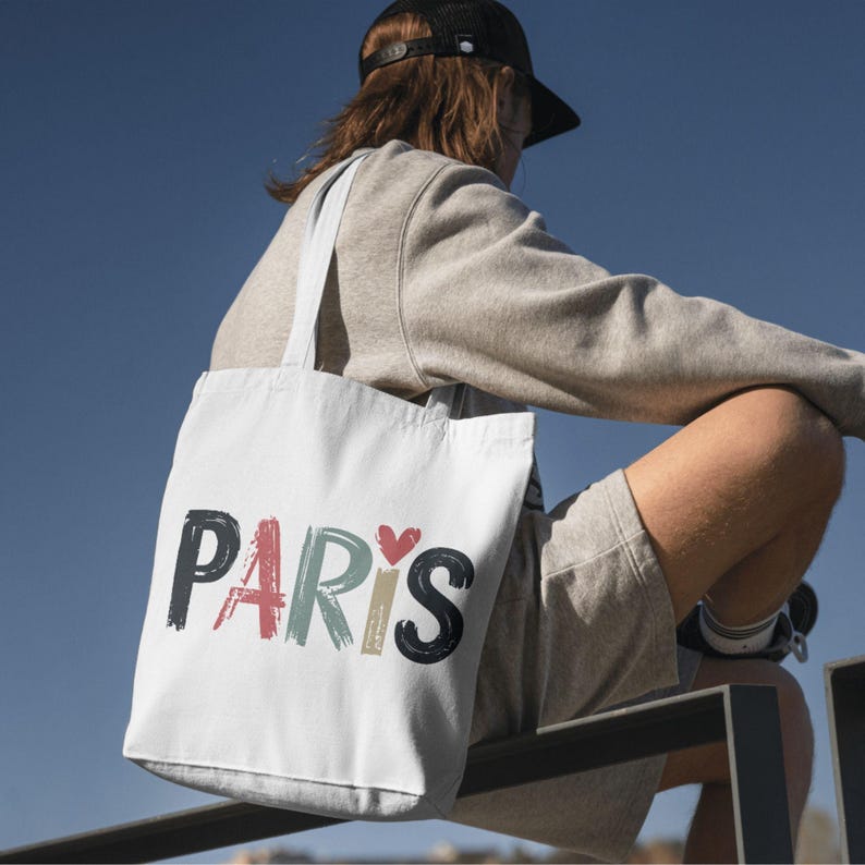 May include: A white tote bag with the word "PARIS" printed in colorful, brushstroke-style lettering. A small red heart is included in the design.