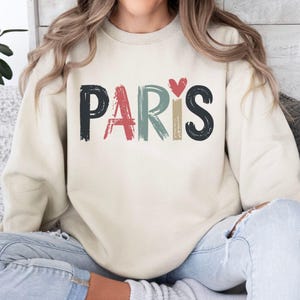May include: A cream colored sweatshirt with the word "PARIS" written in colorful, brushstroke letters. A small red heart is above the "S".