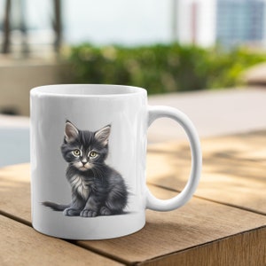 Pu&ograve; includere: Tazza in ceramica bianca con un'illustrazione dettagliata di un gattino grigio e nero con occhi gialli. Il gattino &egrave; seduto, con la coda arricciata. La tazza ha un manico curvo ed &egrave; appoggiata su una superficie di legno.