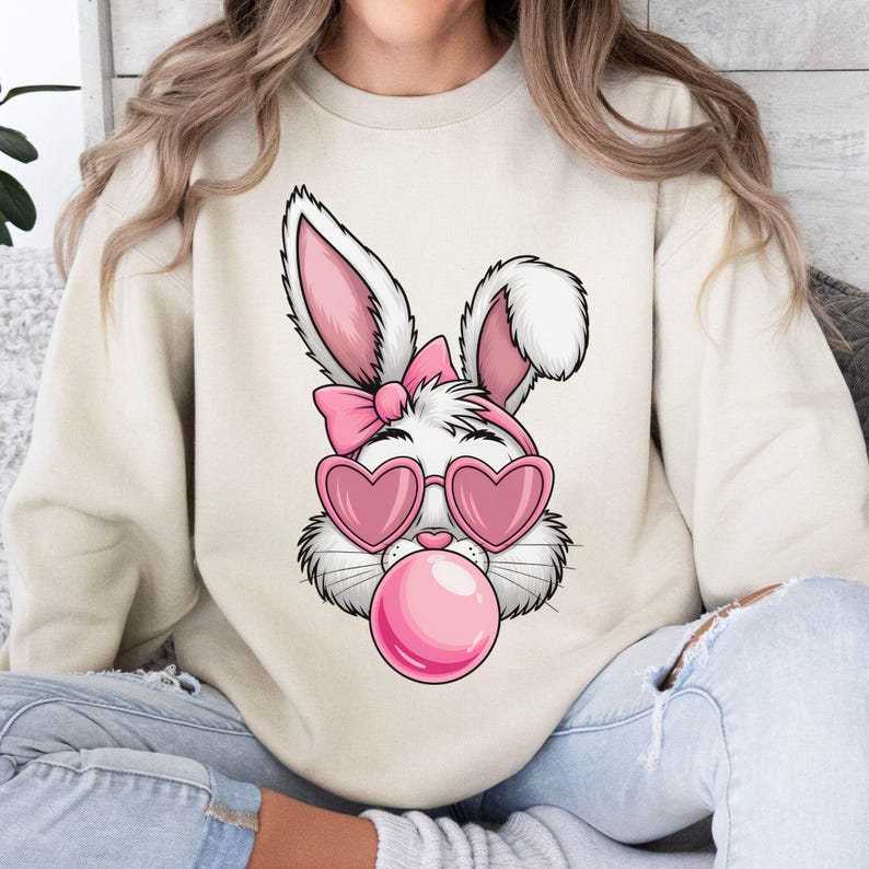 May include: A white bunny wearing pink heart-shaped sunglasses and a pink bow, blowing a pink bubble. The bunny is on a cream-colored sweatshirt.