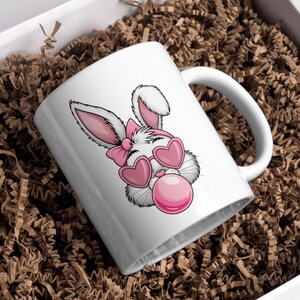 May include: White ceramic mug with a pink and white cartoon bunny wearing heart-shaped sunglasses and blowing a pink bubble.