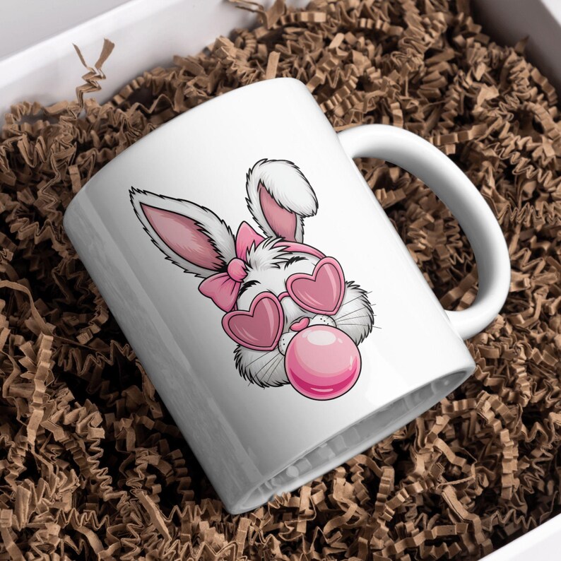 May include: White ceramic mug with a pink and white cartoon bunny wearing heart-shaped sunglasses and blowing a pink bubble.