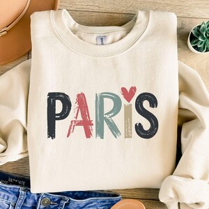 May include: A cream colored sweatshirt with the word "PARIS" written in colorful, brushstroke letters. A small red heart is above the word.