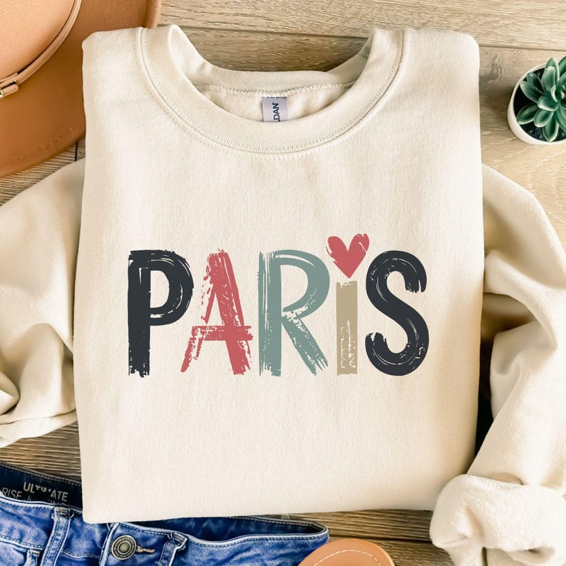 May include: A cream colored sweatshirt with the word "PARIS" written in colorful, brushstroke letters. A small red heart is above the word.