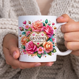 May include: White ceramic mug featuring a floral design with pink and peach roses, green leaves, and the text "BEST GRANDMA EVER" in a heart-shaped frame. The mug is held by a person.