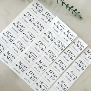 Könnte beinhalten: Ein Bogen mit rechteckigen weißen Aufklebern, die mit schwarzem Text bedruckt sind, der BEYZA ve NURI und das Datum 27.06.2026 zeigt. Die Aufkleber sind in einem Raster angeordnet und tragen jeweils die gleichen Namen und das Datum.
