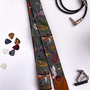 May include: A guitar strap with a dark green background and a pattern of insects and leaves in orange, white, and brown. The strap has brown leather ends. Also pictured are guitar picks, a capo, and a black guitar cable.