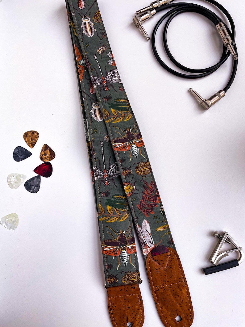 May include: A guitar strap with a dark green background and a pattern of insects and leaves in orange, white, and brown. The strap has brown leather ends. Also pictured are guitar picks, a capo, and a black guitar cable.