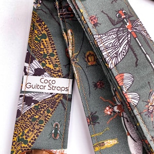 May include: A guitar strap with a repeating insect pattern in shades of green, brown, and orange. The strap features a dragonfly, beetle, and other insect designs. The label reads "Coco Guitar Straps".