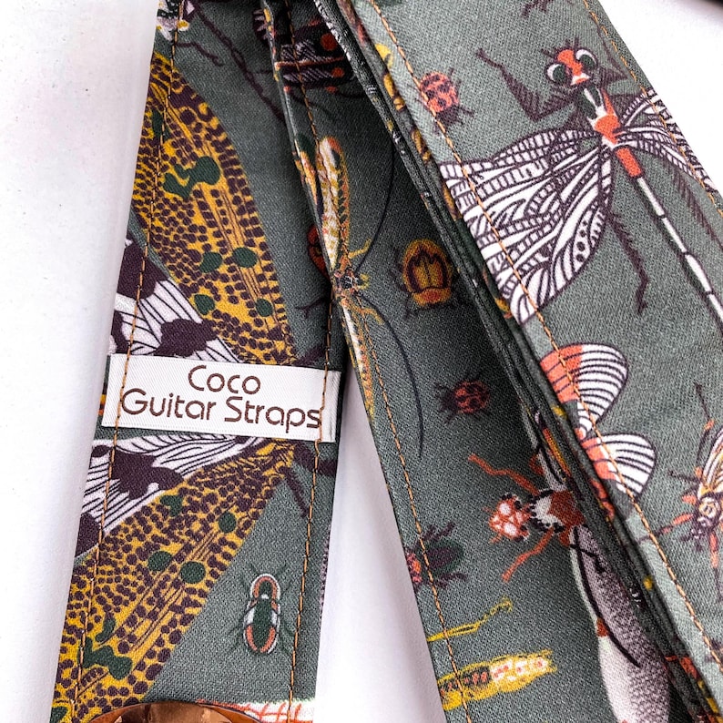 May include: A guitar strap with a repeating insect pattern in shades of green, brown, and orange. The strap features a dragonfly, beetle, and other insect designs. The label reads "Coco Guitar Straps".