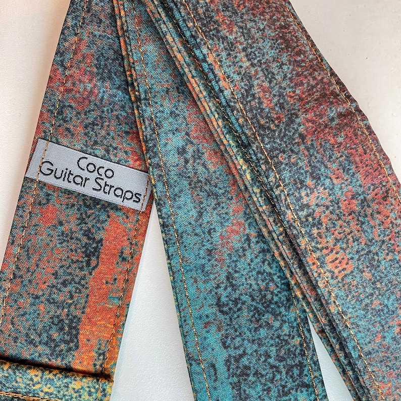 May include: A guitar strap with a blue and orange abstract pattern. The strap is made of fabric and has a label that says "Coco Guitar Straps".