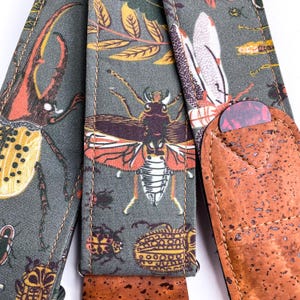 May include: A guitar strap with a gray background and a repeating pattern of colorful insects and leaves. The strap has a brown cork pick holder with a red and purple guitar pick inside.