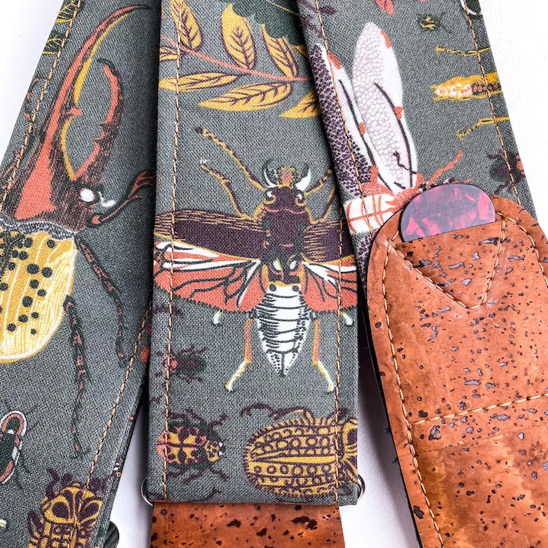 May include: A guitar strap with a gray background and a repeating pattern of colorful insects and leaves. The strap has a brown cork pick holder with a red and purple guitar pick inside.