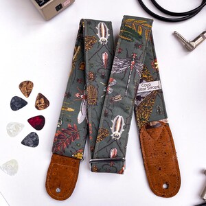 May include: A guitar strap with a dark green background featuring a pattern of insects and leaves. The strap has brown cork ends and a metal buckle. The text "Coco Guitar Straps" is visible on a white label. Guitar picks in various colors are scattered nearby.