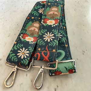 Puede incluir: Dos correas de tela verde oscuro con un dise&ntilde;o floral y de criatura. Las correas tienen herrajes met&aacute;licos plateados y est&aacute;n conectadas entre s&iacute;.