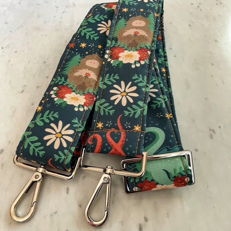 Puede incluir: Dos correas de tela verde oscuro con un dise&ntilde;o floral y de criatura. Las correas tienen herrajes met&aacute;licos plateados y est&aacute;n conectadas entre s&iacute;.