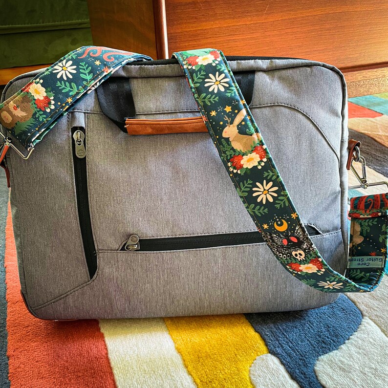Puede incluir: Una bolsa gris para port&aacute;til con una cremallera negra y una correa con estampado floral. La correa tiene un conejo blanco, estrellas y una luna creciente con una calavera y huesos cruzados.