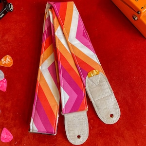 May include: A guitar strap with a colorful geometric pattern in shades of pink, orange, and white. The strap has a white leather end with a cork-like texture.