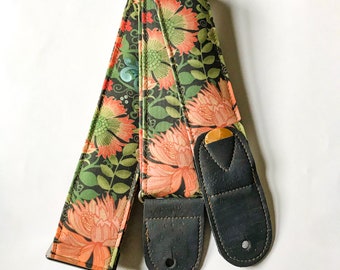 Puede incluir: Una correa de guitarra con un patrón floral en tonos de rosa, verde y negro. La correa tiene dos extremos de cuero negro con una hebilla dorada.
