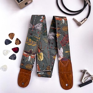 May include: A guitar strap with a dark green background featuring a pattern of insects and leaves in shades of orange, brown, and white. The strap has brown leather ends and a silver metal buckle. Guitar picks and a capo are also visible.